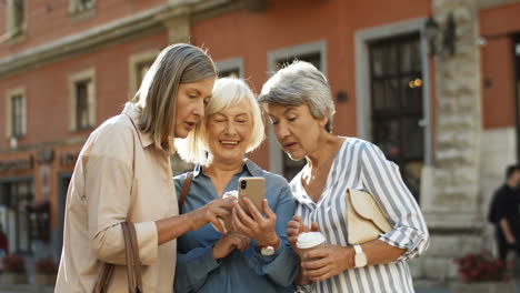 Drei-Schöne-Und-Fröhliche-Frauen-Mit-Kaffee-Zum-Mitnehmen-In-Den-Händen-Lachend,-Während-Sie-Fotos-Auf-Dem-Smartphone-Im-Freien-Scrollen