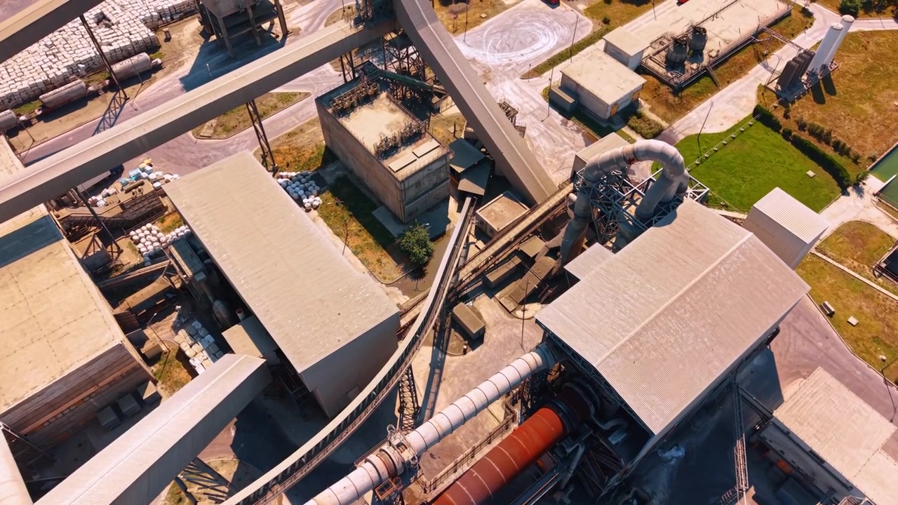 Rotary kiln system and enclosed conveyors from above. Top down aerial view of a red furnace drum, covered belt lines, factory roofs, ducts and concrete service roads.