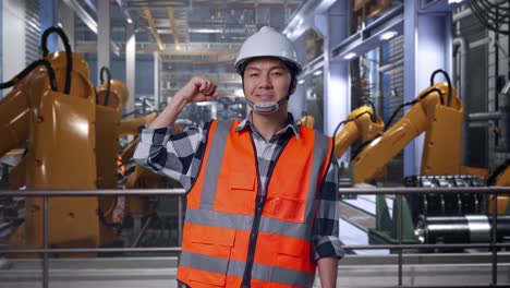 asian male engineer with safety helmet flexing his bicep and smiling to camera while standing with industrial robotic arms working