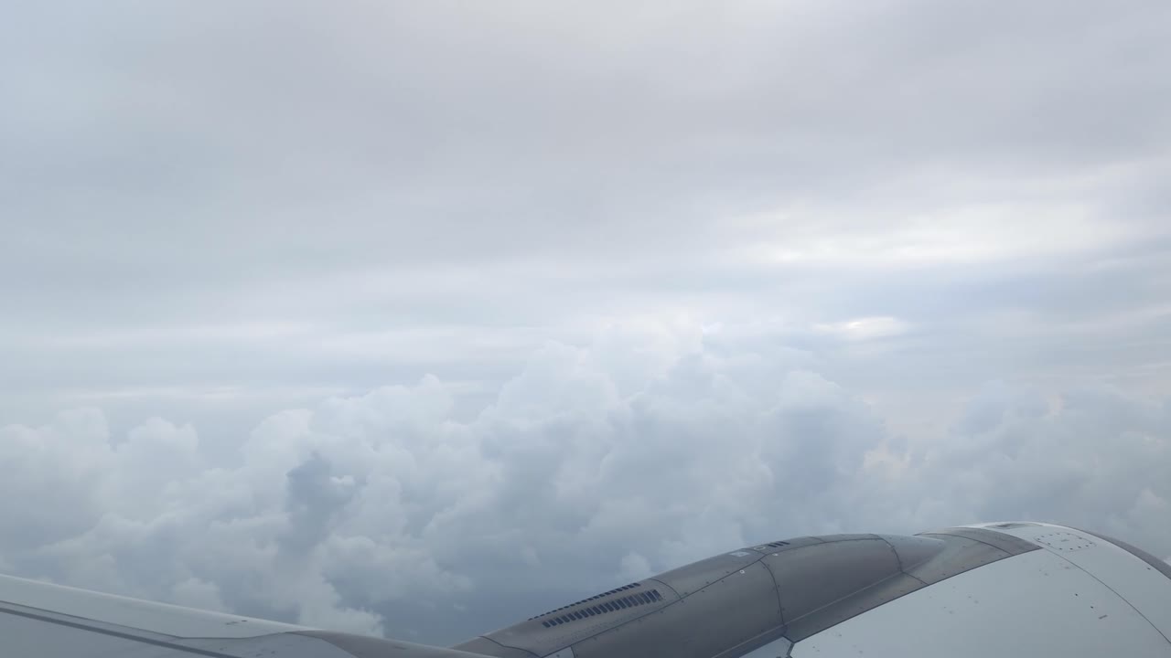 An aerial view from an airplane window reveals the aircraft wing and engine, framed against a dramatic expanse of dense, textured clouds