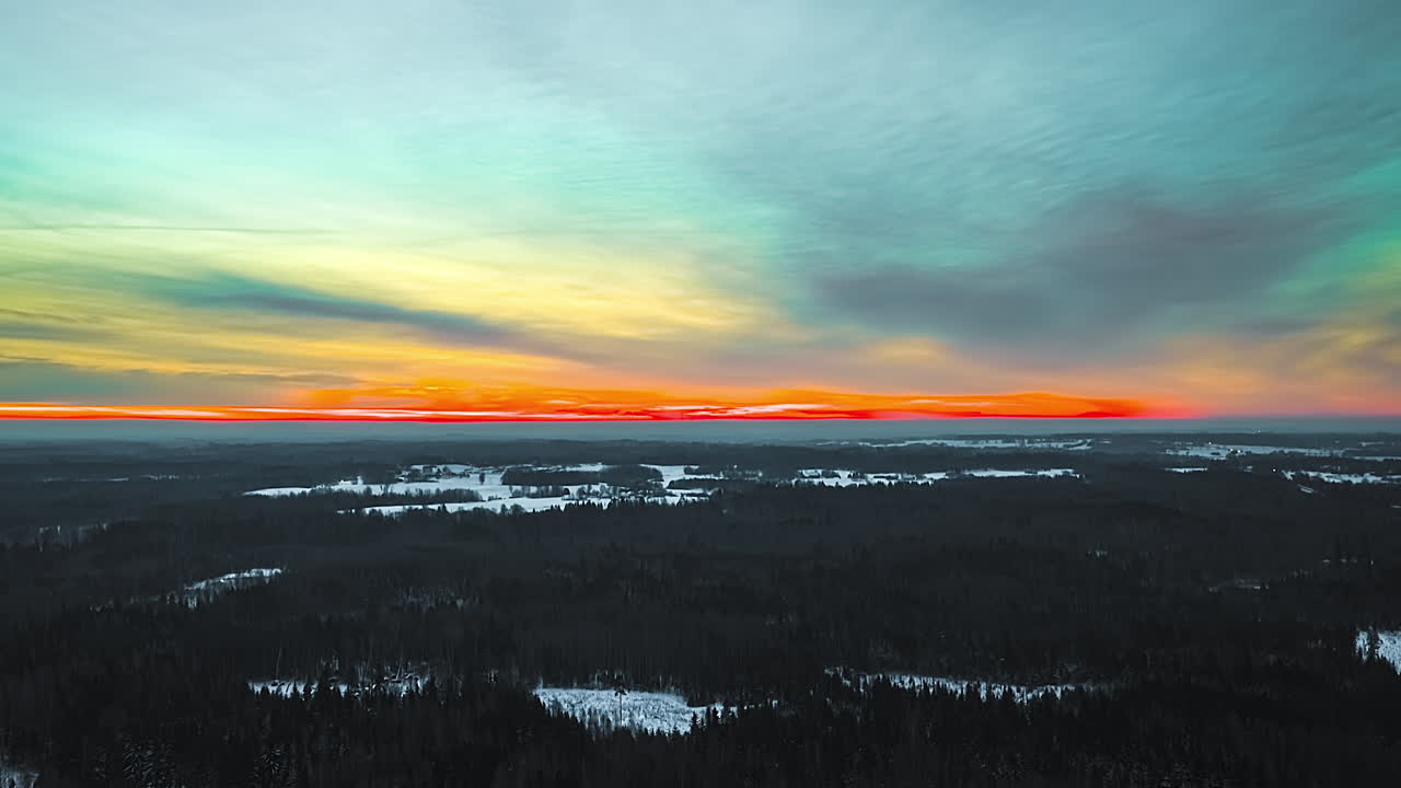 Aerial winter timelapse capturing extraordinary teal and orange post-sunset sky glowing over vast dark pine boreal forest with snow-covered frozen lakes scattered across Latvian wilderness at dusk