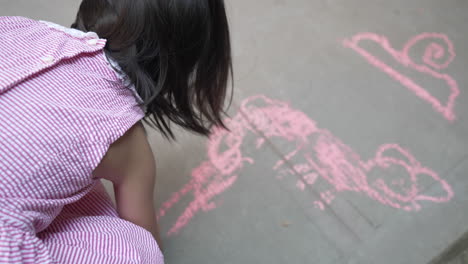 Over-the-shoulder-view-of-a-girl-in-dress-drawing-with-a-pink-chalk-on-concrete-ground
