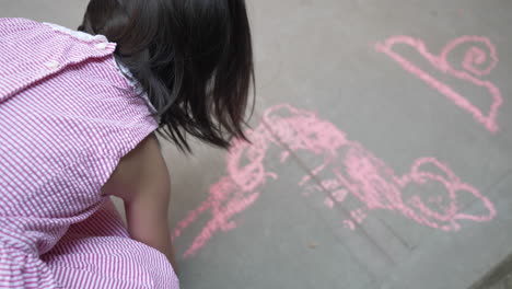 blick über die schulter eines mädchens im kleid, das mit rosa kreide auf betonboden zeichnet