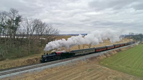 una vista de drones de una locomotora de vapor con vagones de pasajeros acercándose con una cabeza llena de vapor sobre el campo en un día de invierno