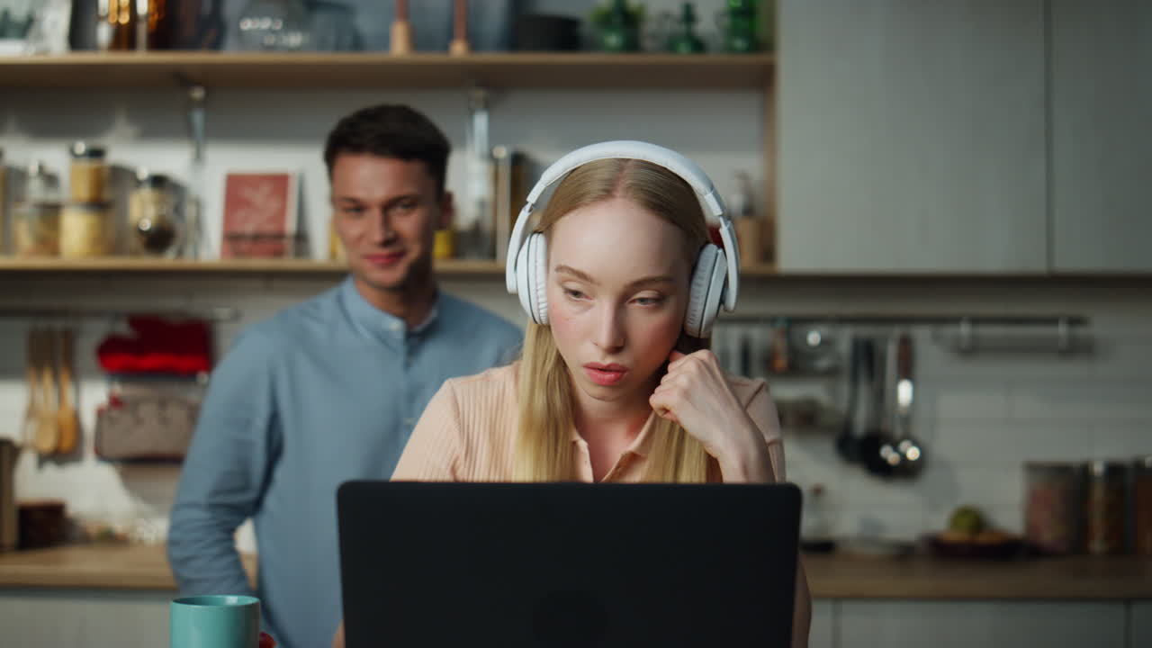 Woman watching internet video laptop by headphones at kitchen with man behind.