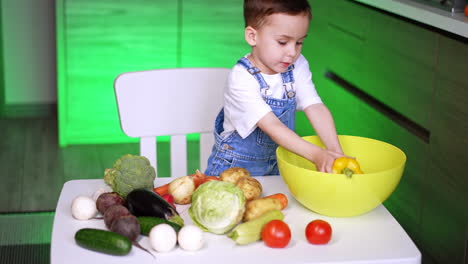 caucasian toddler in white t-shirt and jeans romper sits at table. little kid learns the vegetables in the kitchen.