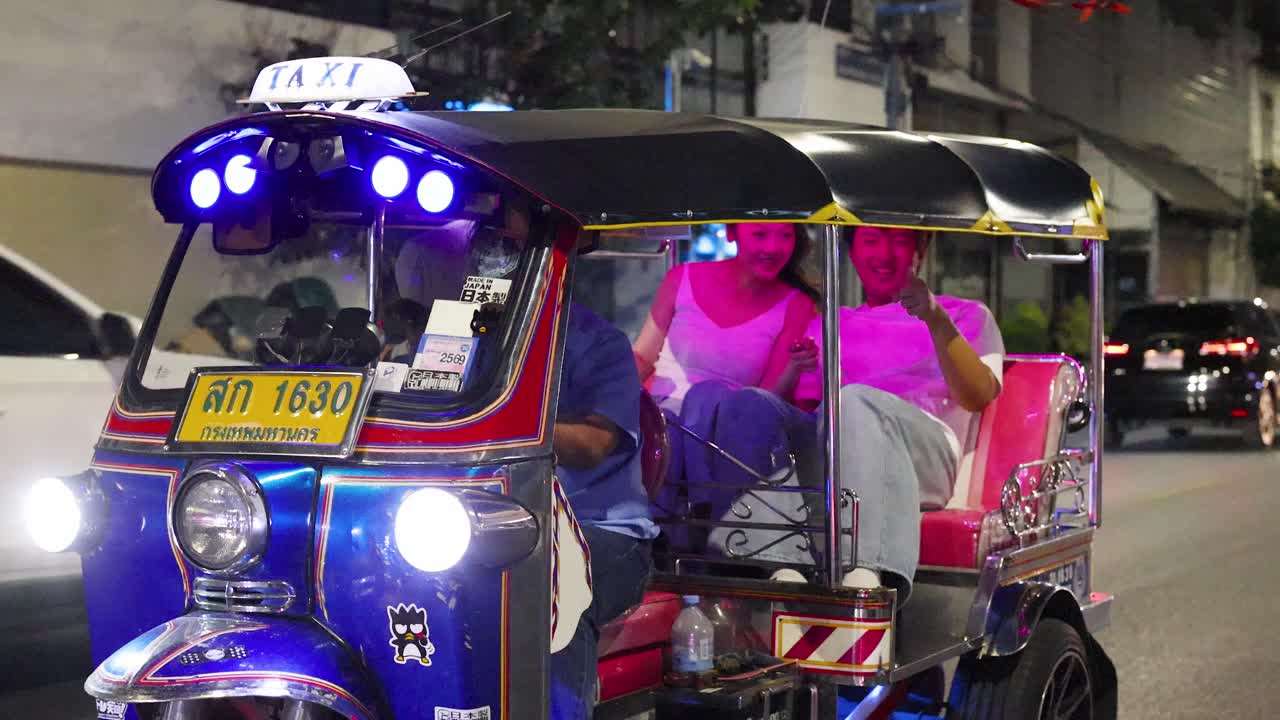 Un couple de touristes à bord d'un tuk-tuk taxi illuminé sur une rue urbaine animée la nuit, sous des lumières vibrantes.