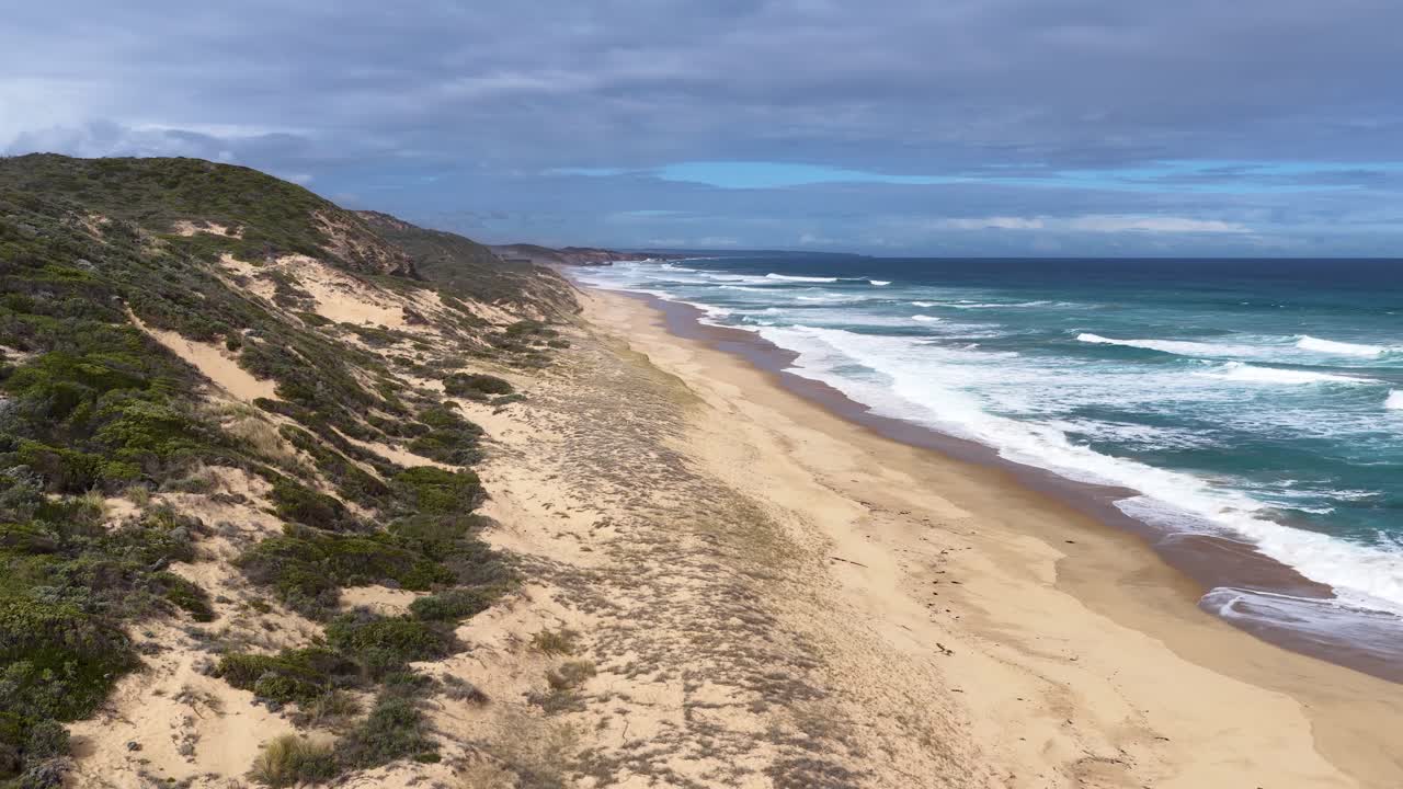 Drone glides above sandy coastline, green cliffs, and rolling waves under bright, partly cloudy sky
