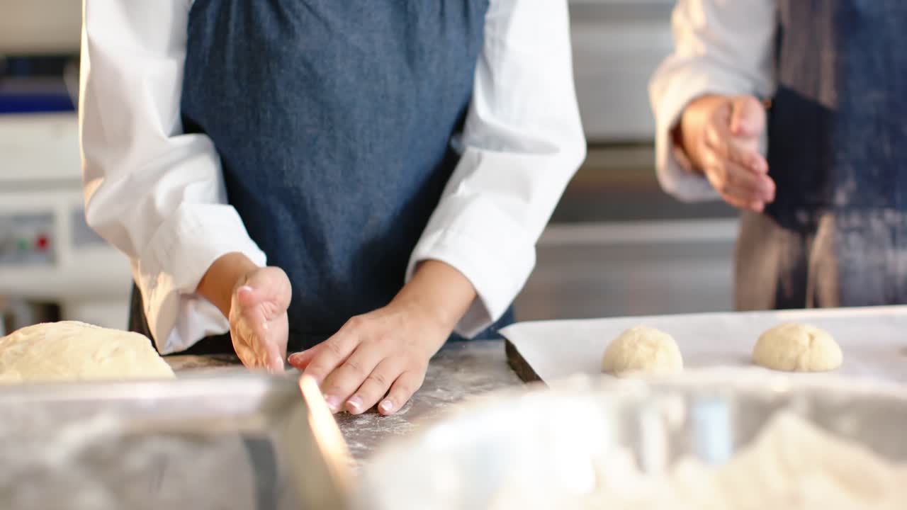 Diverse female bakers shaping buns after portioning at prep counter with tray in aprons for baking