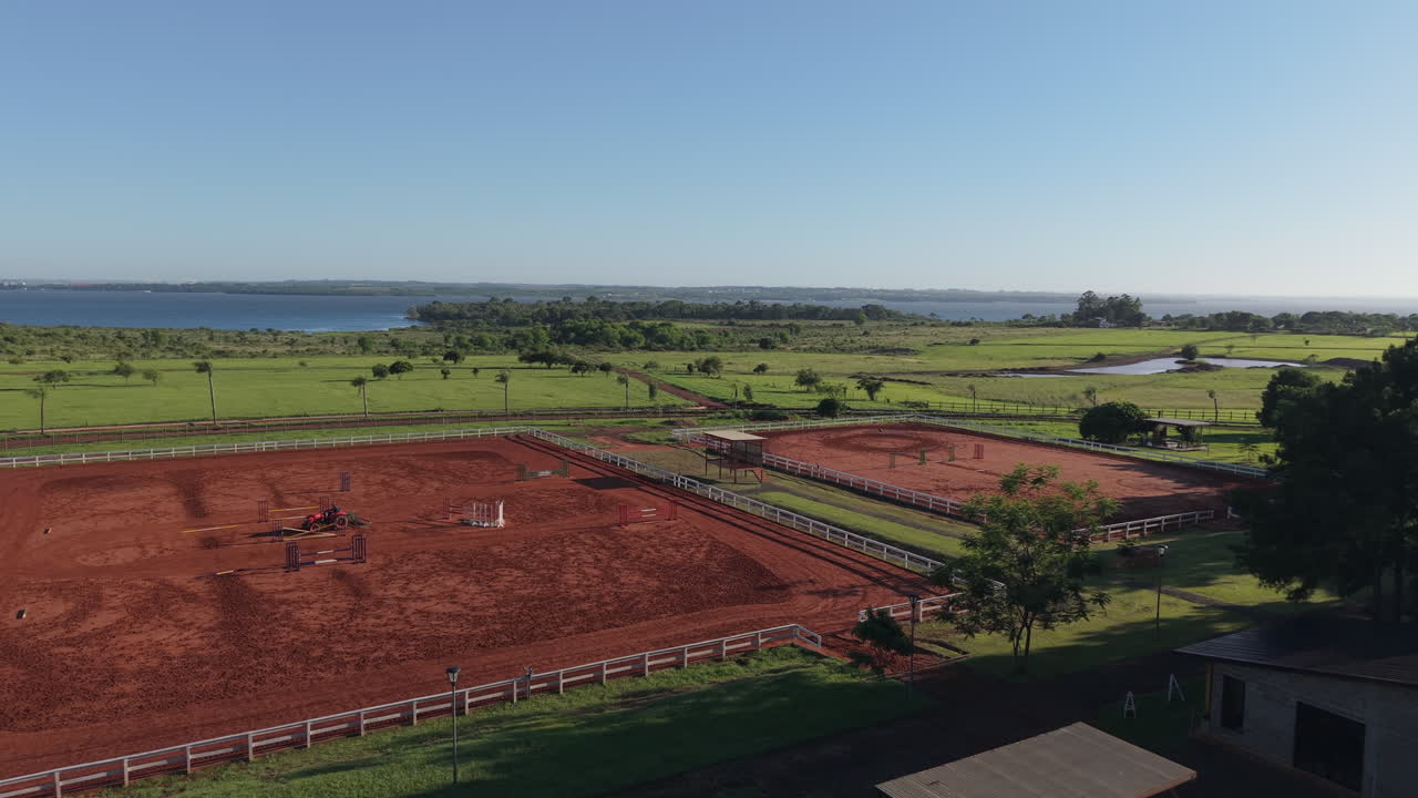 Wide-angle aerial view of horse riding and training ground with red dirt arenas and open rural landscape near the waterside.