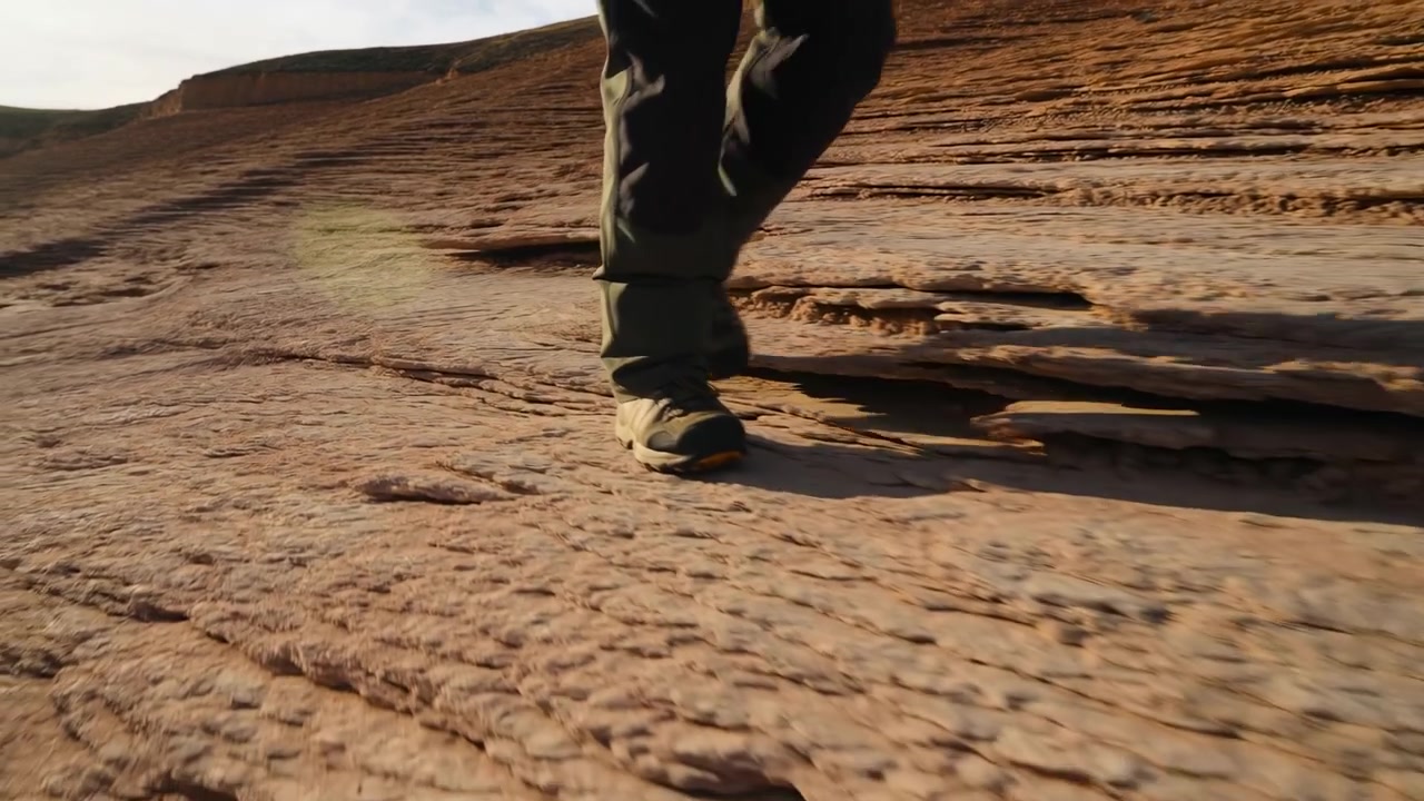 Scena ravvicinata dalla vita in giù di una persona che cammina su una sporgenza di arenaria stratificata, mostrando pantaloni outdoor color oliva, scarpe da trekking robuste, una superficie rocciosa texturizzata, luce solare calda e un ambiente di sentiero desertico all'aperto.