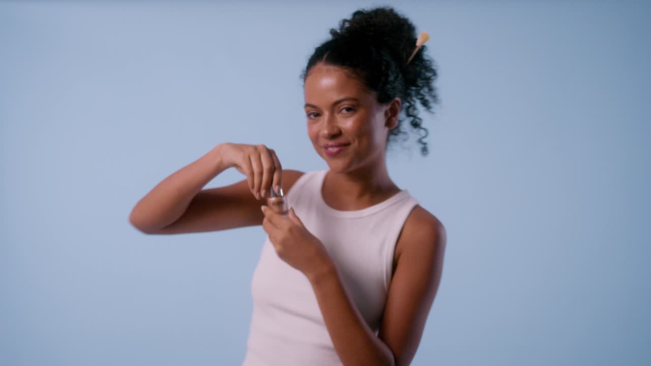 A young model uses a brush to apply facial product from her hand, smiles, and playfully touches her face with the brush.