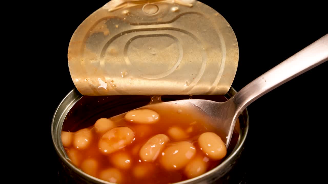 A metal spoon lifts baked beans in tomato sauce from an open can against a black background, with close-up, well-lit, steady shots