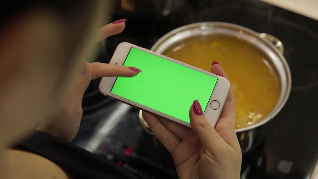 mujer está sosteniendo un teléfono inteligente con una pantalla verde, y cocinando sopa