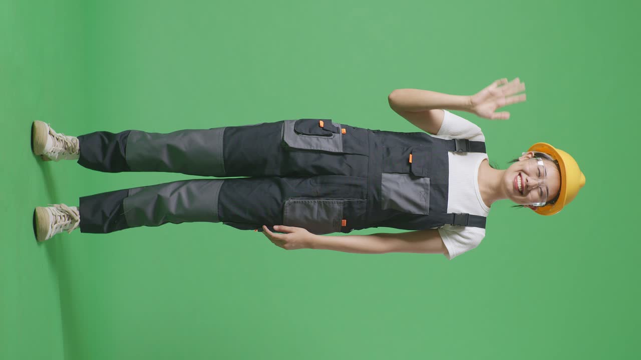 안경과 안전 헬을 착용한 아시아 여성 노동자가 미소 짓고 손을 흔들며 녹색 스크린 배경 스튜디오에 서서 작별 인사를 하고 있습니다.