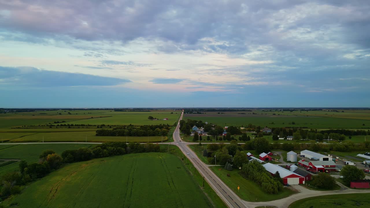 Una vasta veduta aerea cattura un tranquillo paesaggio rurale al crepuscolo, con una piccola fattoria con fienili rossi e silos bianchi immersi tra vivaci campi verdi.