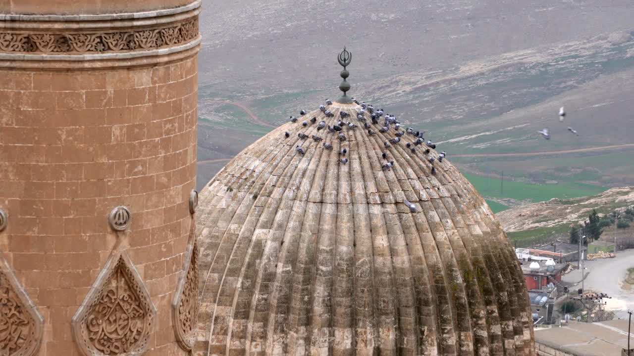 el minarete y la cúpula de mardin ulu camii en el mismo marco, la cámara se está acercando a la cúpula donde se pueden ver las palomas posándose en la cúpula en un día nublado