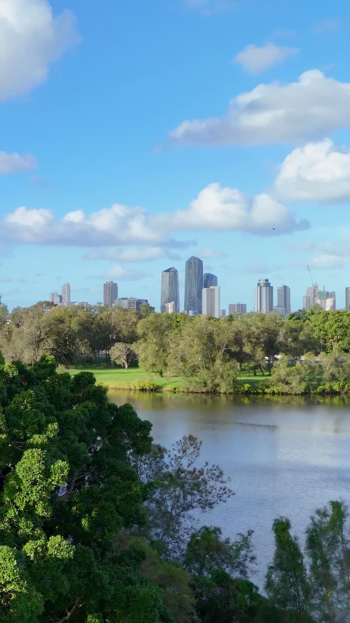 Uma cena de parque serena com vegetação exuberante e um rio, tendo como pano de fundo o horizonte da cidade de Gold Coast sob um céu azul brilhante.