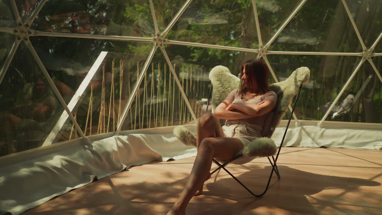 mujer pensativa con las manos cruzadas se sienta en el sillón. la dama se relaja en la ventana panorámica de la terraza. la hembra disfruta de la vista del paisaje forestal en una atmósfera tranquila