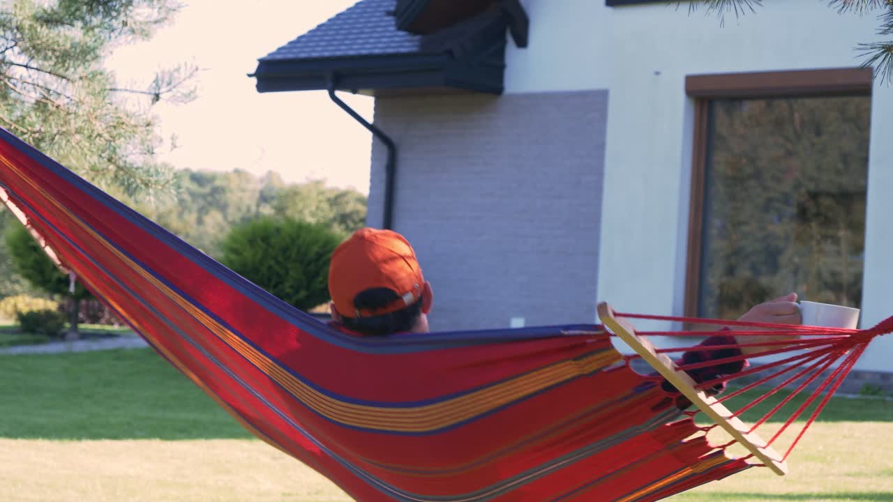 Man Swinging In A Hammock While Drinking A Hot Coffee In The Morning. - wide shot