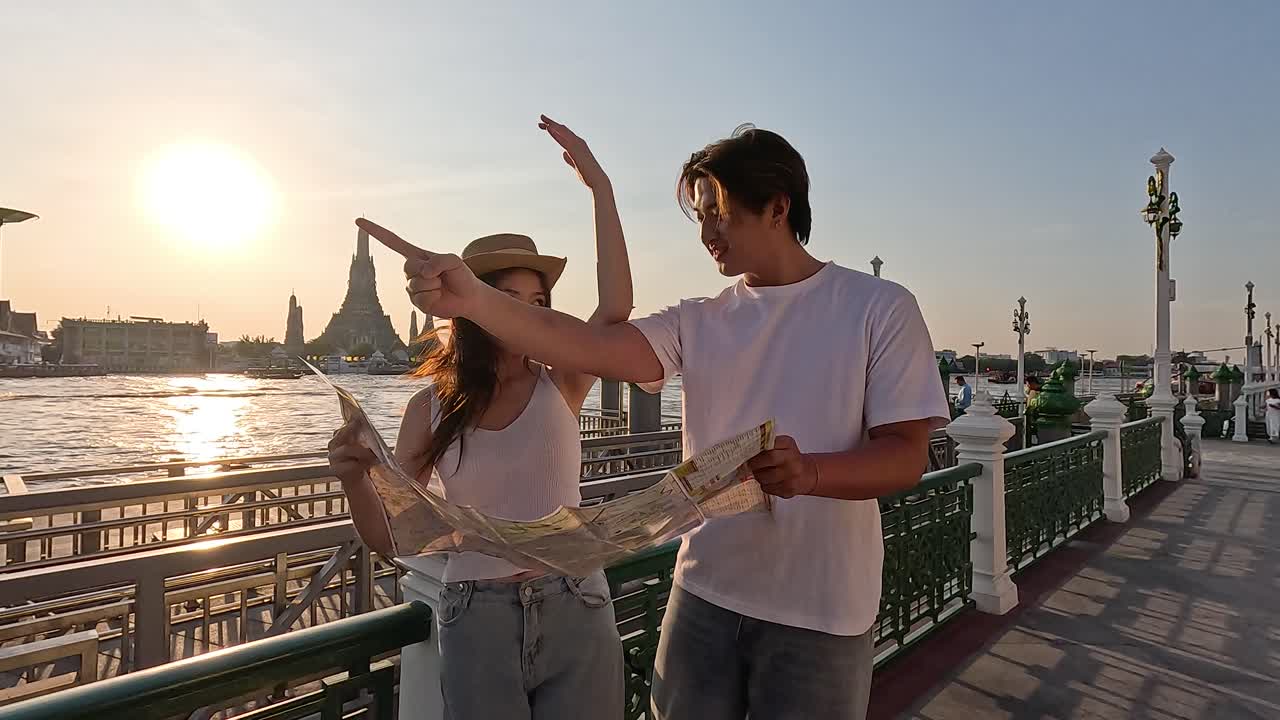 Coppia di turisti studia una mappa cartacea su un lungofiume al tramonto, con controluce caldo e telecamera fissa.