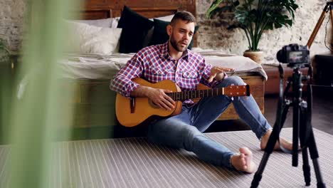 professional musician blogger is recording tutorial about playing guitar for internet blog using camera. he is talking to followers showing instrument then playing and singing.