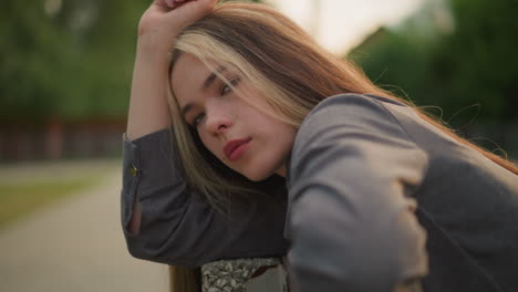 close-up of lady in grey clothing resting her head on arm outdoors, lost in thought with a calm expression, she blinks softly, while the background remains blurred