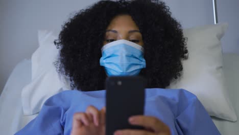 Afroamerikanische-Patientin-Mit-Gesichtsmaske,-Die-Auf-Dem-Krankenhausbett-Liegt-Und-Ihr-Smartphone-Benutzt