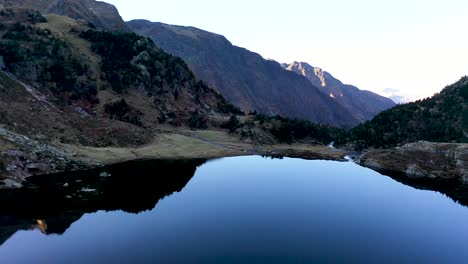 Lac-d'Espingo-reflecting-mountain-lake-located-in-Haute-Garonne,-Pyrénées,-France,-Aerial-flyover-pan-right-shot