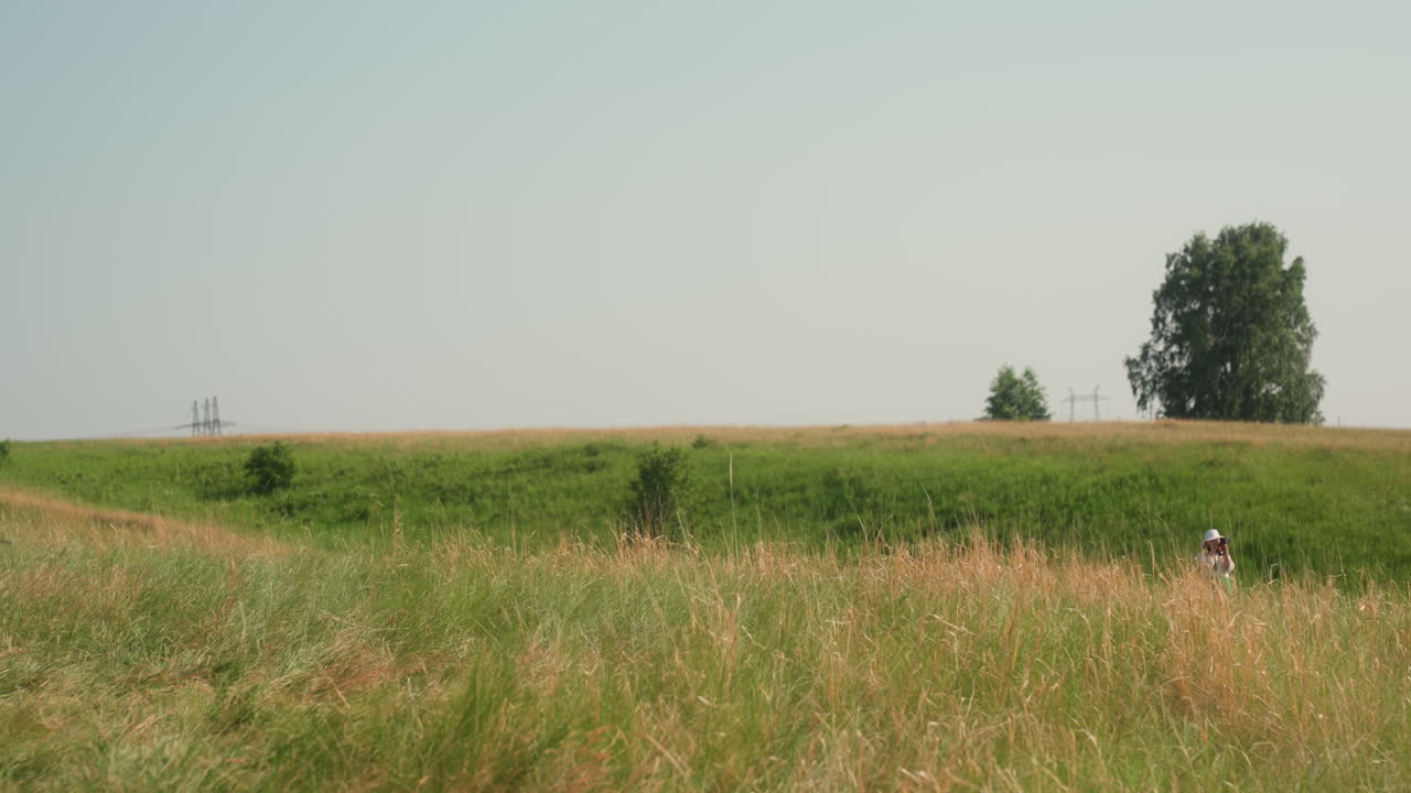 Woman in white hat walks through distant tall grass surrounded by lush green field, soft golden tones, and calm sky, creating peaceful rural atmosphere with trees and power lines in background