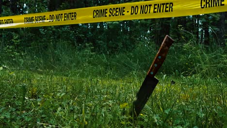 low angle close up footage of a bloody knife standing upright in grassy ground in front of a yellow police crime scene do not enter tape indicating a crime has been commited and investigation happens