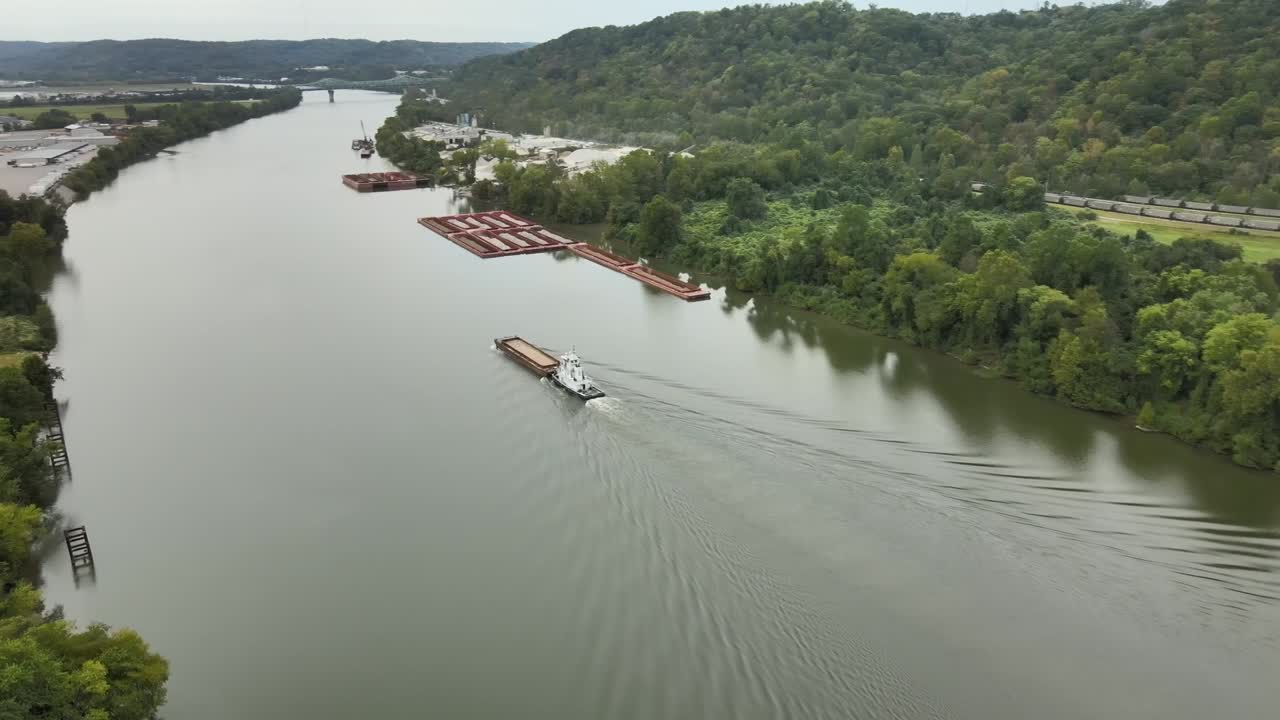 Aerial drone footage of towboat guiding cargo barge along calm Kanawha River surrounded by forested hills, industrial docks, and quiet roads in scenic West Virginia, United States