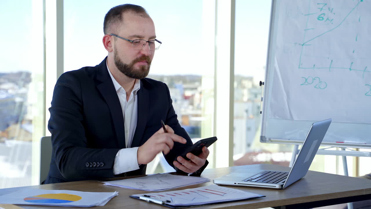 Bearded businessman in glasses working in office. Serious office worker in suit sitting at the desk using the phone and talking.