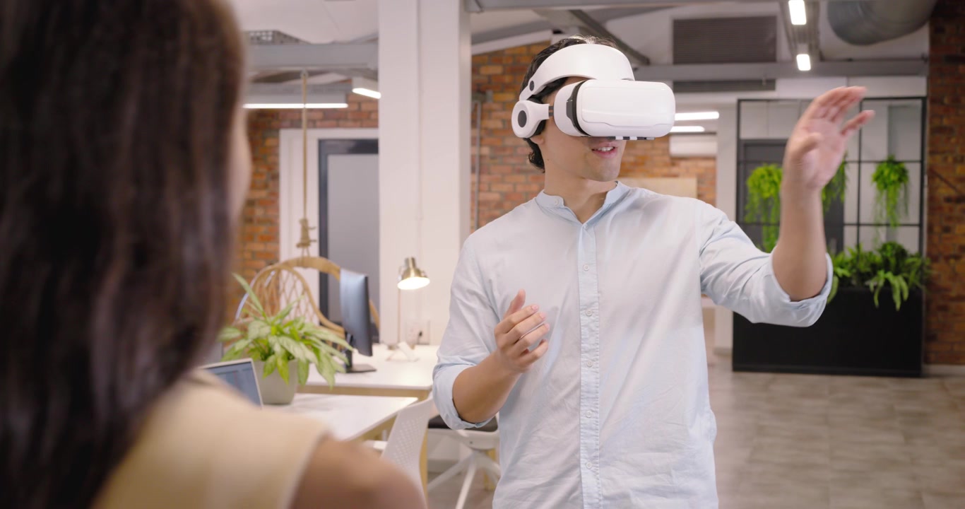 Team d'ufficio eterogeneo che dimostra la VR, uomo asiatico che indossa un visore VR bianco che si protende mentre una donna lo guida