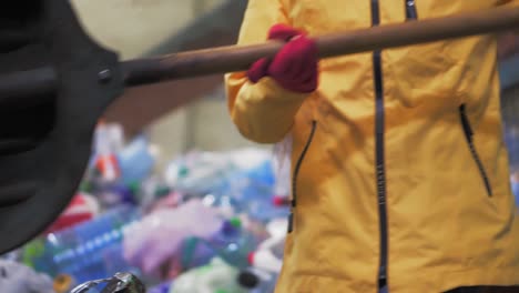 Nahaufnahmen-Einer-Person-In-Gelber-Jacke-Und-Handschuhen,-Die-In-Einer-Kunststoffrecyclingfabrik-Gebrauchte-Flaschen-Mit-Einer-Großen-Entenschaufel-Schaufelt.-Riesiger-Stapel-Flaschen-Im-Hintergrund