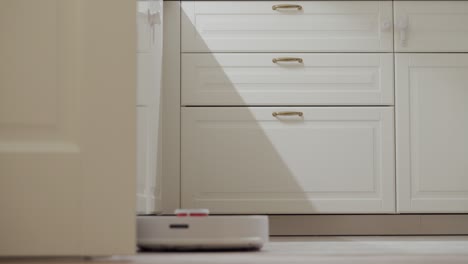 white robot vacuum cleaner cleaning floor in the kitchen white room interior in the evening.