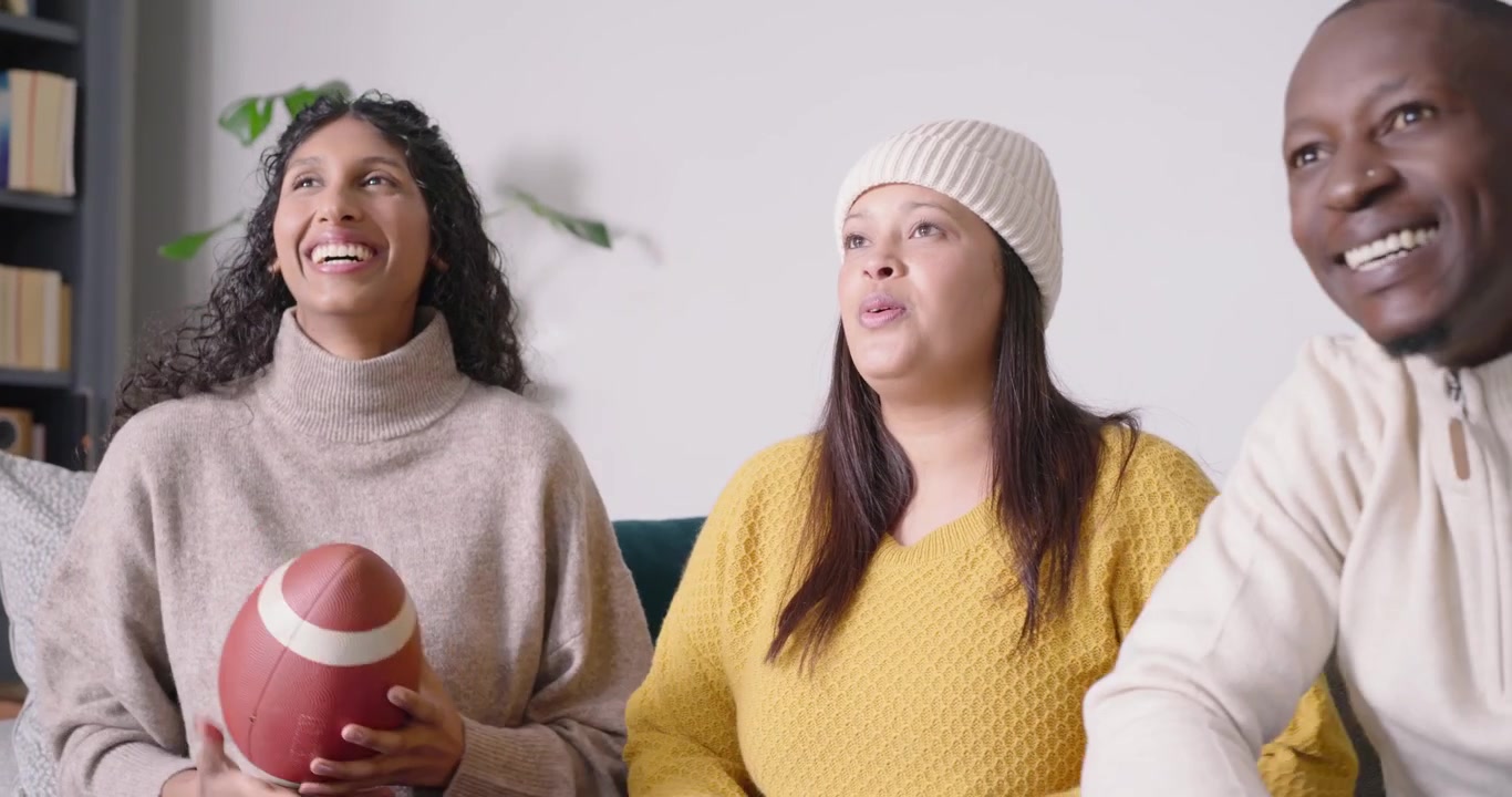 Amici afroamericani seduti sul divano a casa che reagiscono a un gioco in TV, passandosi un pallone da football americano, esultando.