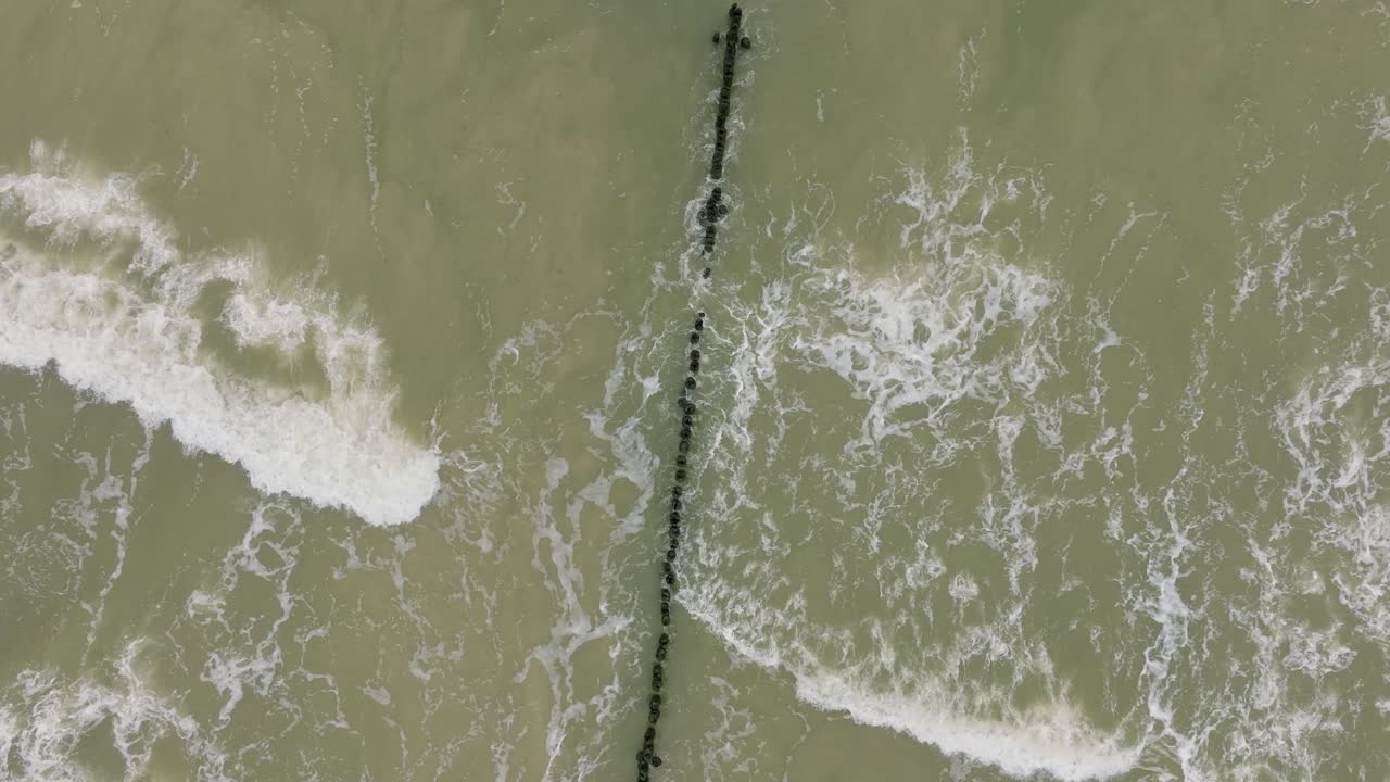 Aerial birdseye view of Baltic sea coast on a overcast day, old wooden pier, white sand beach, large storm waves crushing against the coast, climate changes, wide drone orbit shot