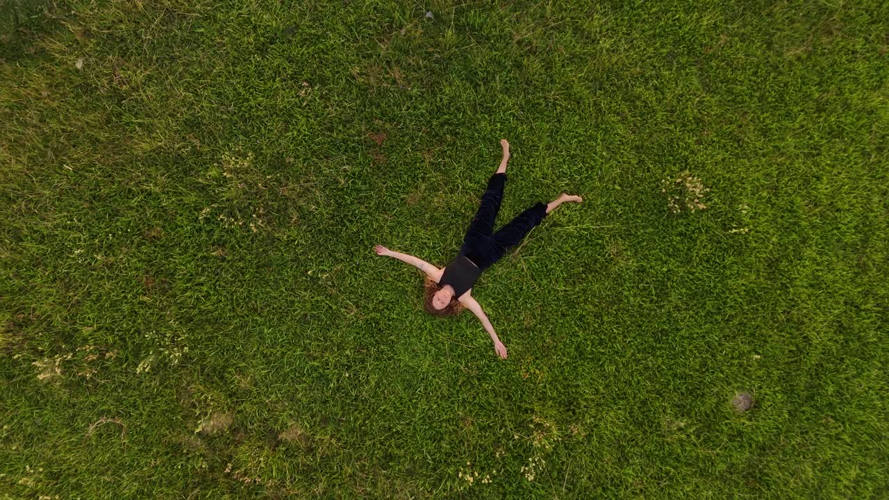 Aerial top-down shot descending slowly towards a woman lying peacefully on her back in lush green grass, arms outstretched, relaxing in nature