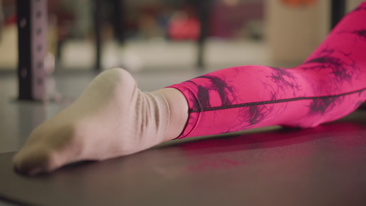 donna bianca che allunga piede e polpaccio sul tappetino, primo piano sulla mobilità della caviglia e delicata dorsiflessione della caviglia, atmosfera da paziente in riabilitazione e studente di pilates, leggings a fantasia rosa con calzini morbidi, studio a bassa angolazione