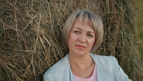 middle-aged woman rests thoughtfully on hay bale in vast farmland, she gazes into the distance, surrounded by rolling fields, creating a peaceful and reflective rural moment under an open sky