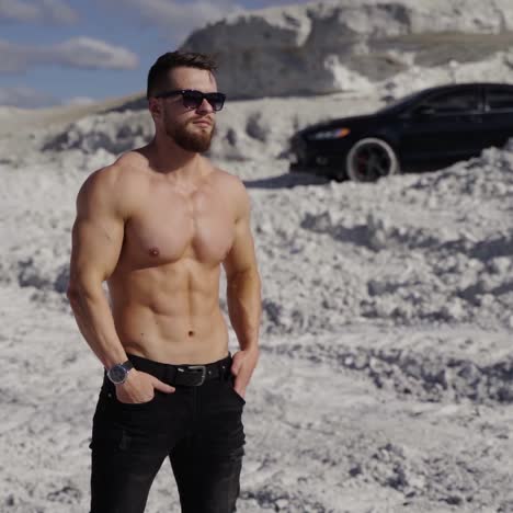 athlete with muscular body in white mountains. handsome sportsman without shirt in sunglasses stands on canyon on black car background.