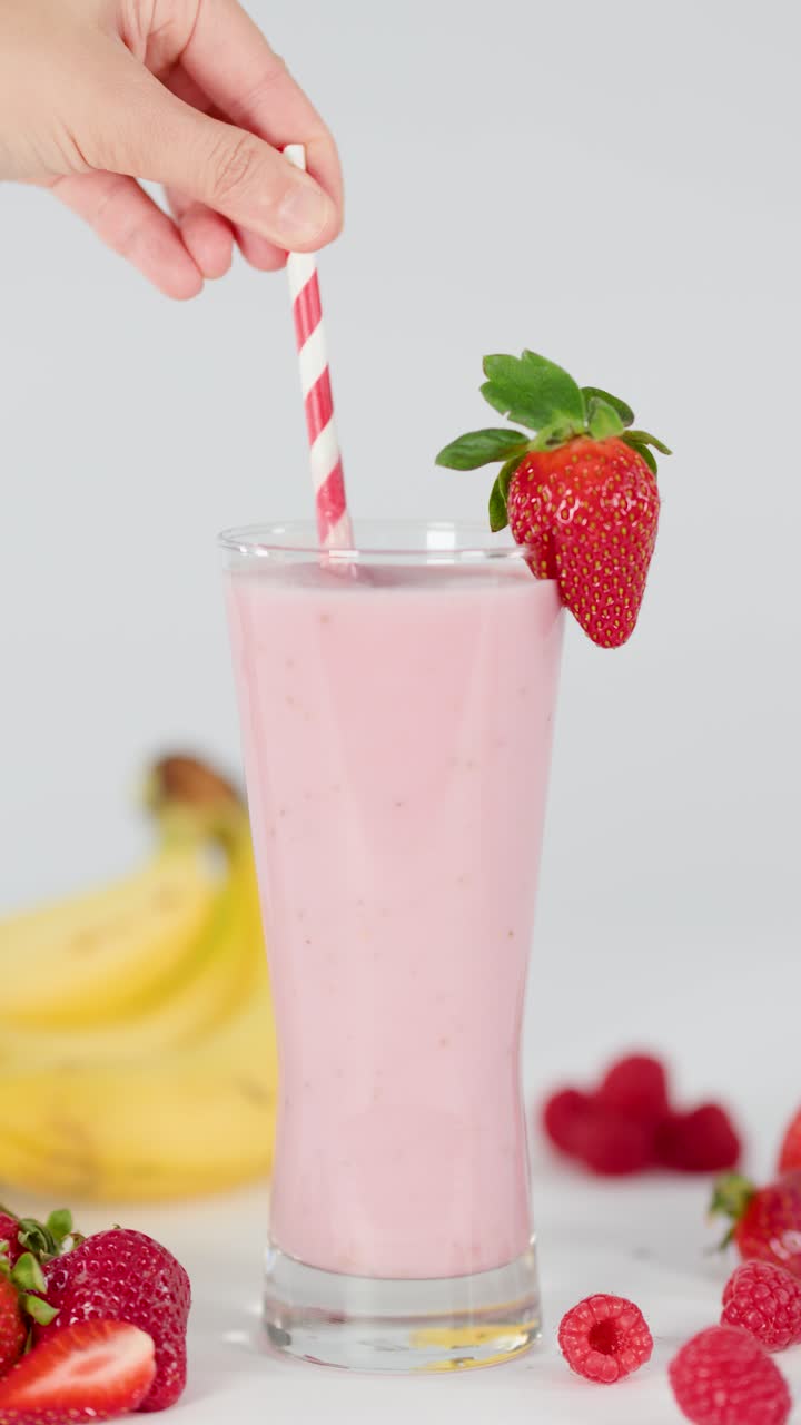 Una mano revuelve un batido rosa cremoso con una pajita a rayas, rodeado de fruta fresca, con iluminación brillante.