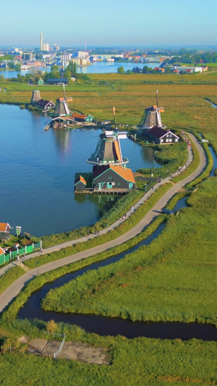 Expansive aerial view of the iconic windmills and traditional houses of Zaanse Schans. A modern city skyline looms in the distance beyond green fields and winding waterways, showing contrast.