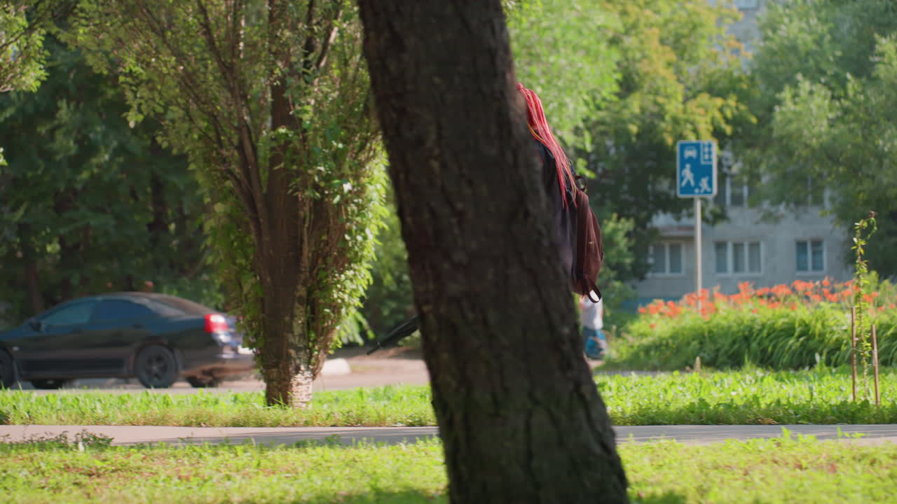 찰랑이는 땋은 머리를 한 Parkside의 여성, 번잡한 거리 가장자리를 걷는 캐주얼한 여성, 낮 동안 길가의 차량을 지나면서 네온 땋은 머리를 보여주는 편안한 검은색 복장의 여성