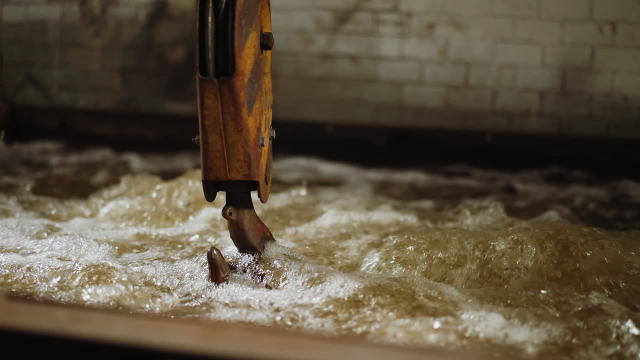 Illuminated by subtle lighting, a crane hook circulates through bubbling water in an industrial quench tank, causing a steamy and dynamic water surface