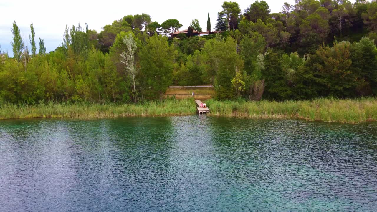 Ripresa aerea in allontanamento da una coppia seduta su una piattaforma di legno davanti a un lago con acqua limpida, tra gli alberi e una casa sullo sfondo, a Banyoles, Catalogna, Spagna