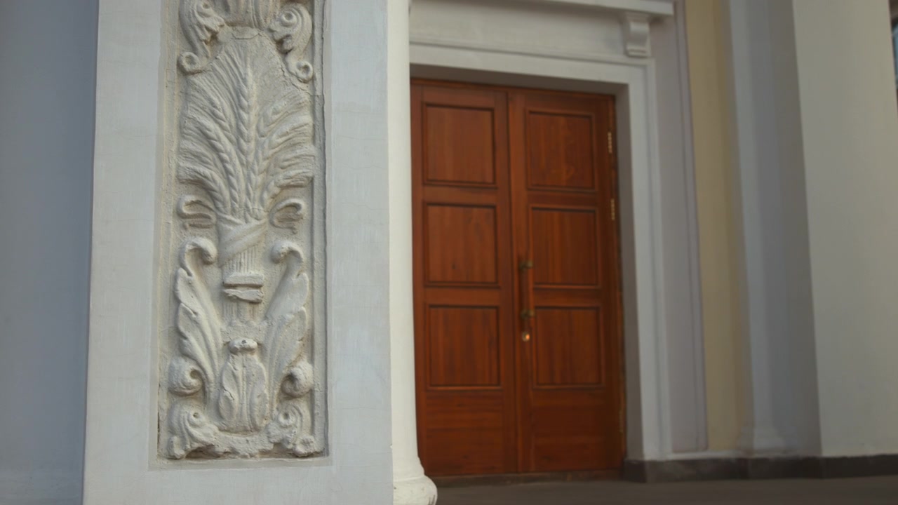 Vista a livello degli occhi di un ingresso architettonico ornato che mostra un pilastro in pietra scolpita con rilievo a foglie d'acanto in primo piano a sinistra e un'alta porta doppia in legno incastonata in un portale incassato, con luce soffusa e ricca