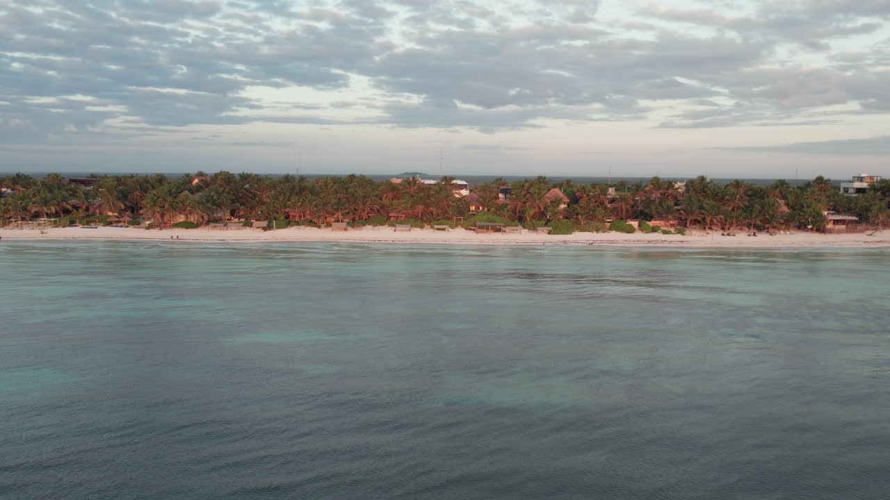 tomada aérea de cabañas y chozas rodeadas de palmeras frente a una playa de arena blanca con un océano azul cristalino en tulum, méxico