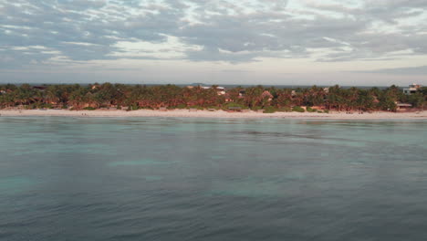 Aerial-pull-back-shot-of-cabins-and-huts-surrounded-by-palm-trees-in-front-of-a-white-sand-beach-with-a-crystal-clear-blue-ocean-in-Tulum,-Mexico